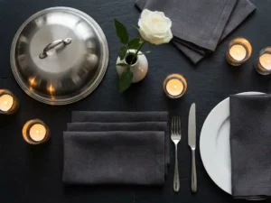 Zilveren serveerdomes, witte roos en gevouwen linnen servetten op leisteen, elegante tafelsetting met kaarslicht en fijn porselein.