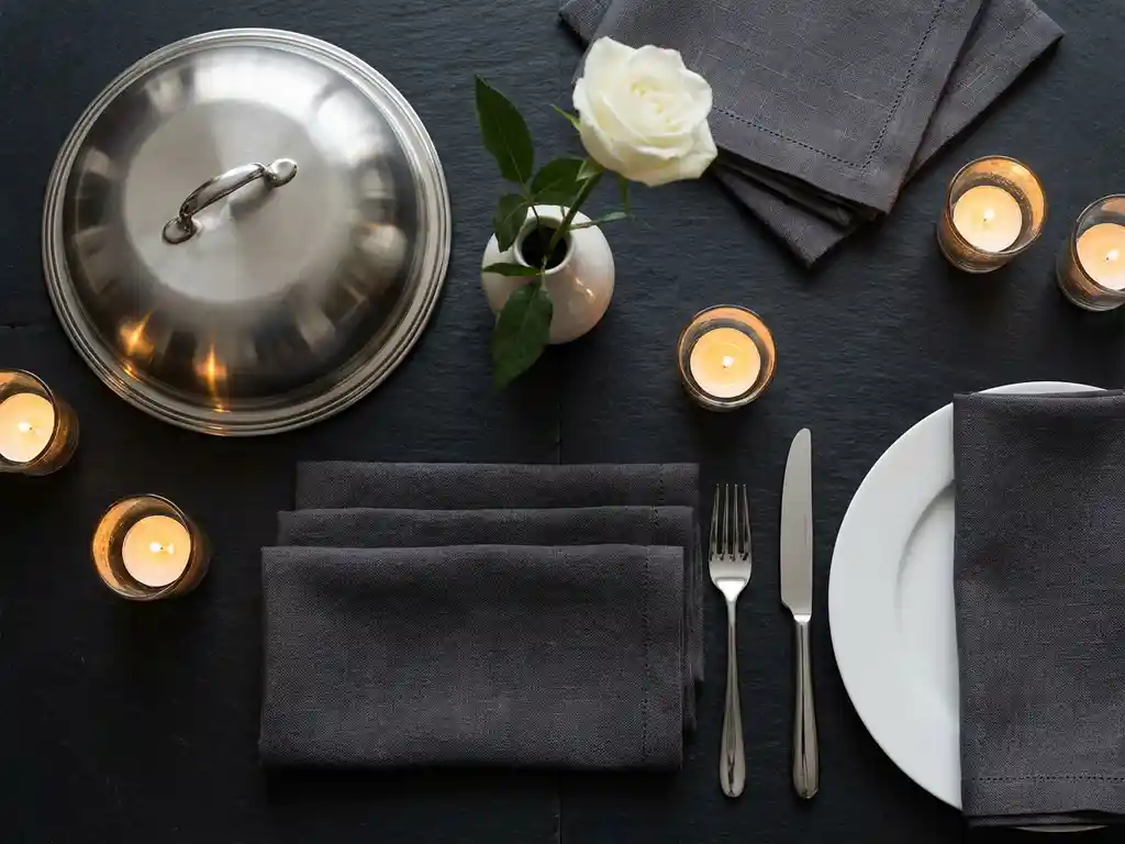 Zilveren serveerdomes, witte roos en gevouwen linnen servetten op leisteen, elegante tafelsetting met kaarslicht en fijn porselein.