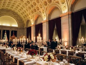 Historische museumzaal ingericht als zakelijke eventlocatie met witte linnen tafels, kristallen glaswerk en kaarslicht onder vergulde plafonds.