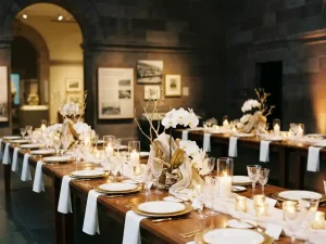 Feestelijk gedekte bankettafels met witte linnen, sculpturale bloemstukken en kristallen glaswerk in een sfeervolle museumzaal.
